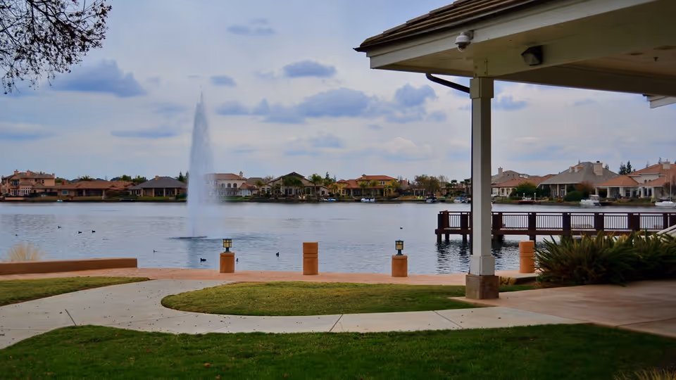 View of a peaceful lake with a water fountain in the center, surrounded by houses on the far shore. In the foreground, there is a covered patio area with a wooden deck extending over the water, some green grass, and a paved walkway.