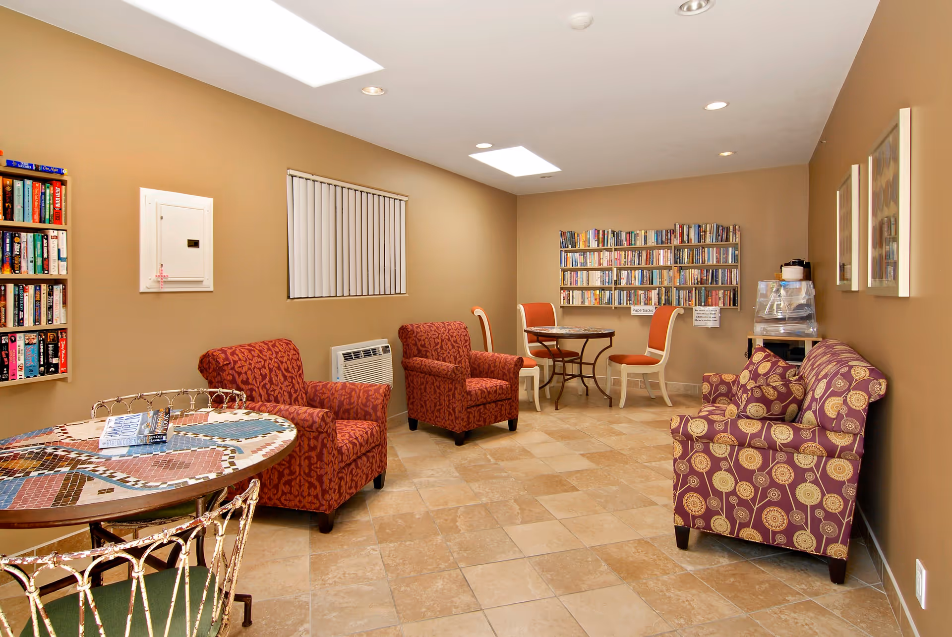 A cozy senior living common area with beige walls and tiled floor, featuring two red patterned armchairs, a purple patterned armchair, two small round tables with chairs, and bookshelves filled with books mounted on the walls.