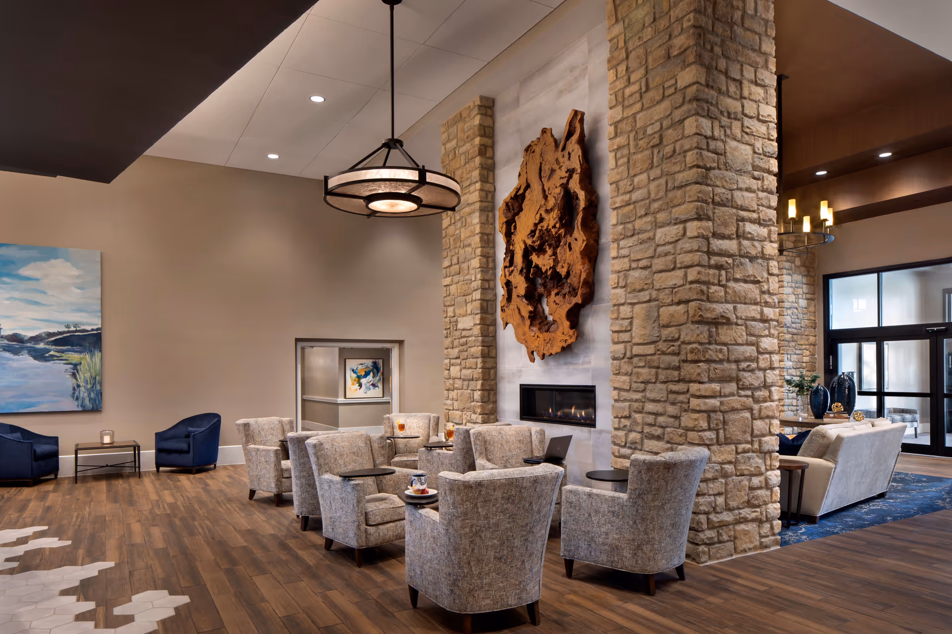 A spacious and elegant common area with a stone fireplace as the focal point, surrounded by comfortable upholstered chairs and small tables. The room features wooden flooring with a unique hexagonal tile pattern, modern pendant lighting, and a large abstract wooden wall art above the fireplace. There are additional seating areas with sofas and armchairs, and large windows near the entrance letting in natural light.