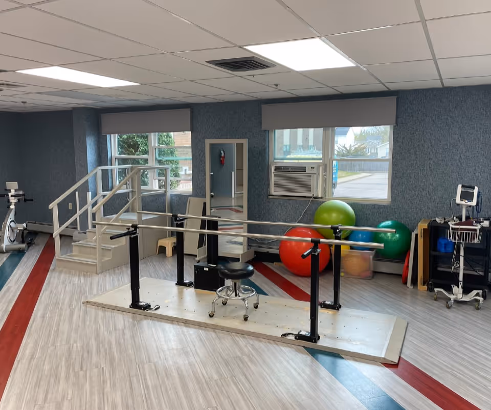 A physical therapy room with parallel bars for walking exercises, a small set of stairs with handrails, exercise balls in various colors, a stationary bike, a mirror, and medical equipment on a cart. The room has large windows with an air conditioning unit and light-colored flooring with red and blue stripes.