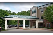 Exterior view of a multi-story nursing and rehabilitation facility with a covered entrance supported by white columns. Several cars are parked in the parking lot, and trees surround the building.