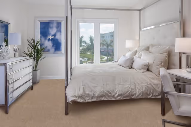 Bright and modern bedroom with a large bed featuring a tufted headboard and multiple pillows. The room has a mirrored dresser with a lamp and decorative items, a potted plant, and a framed picture of a bird in the sky on the wall. Large windows provide a view of palm trees and buildings outside.