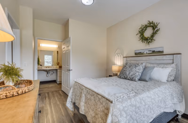 A cozy bedroom with a neatly made bed featuring patterned bedding and multiple pillows. A bedside table with a lamp is next to the bed. On the wall above the bed is a decorative wreath and a small landscape picture. The room has light-colored walls and wood flooring. An open door reveals a bathroom with a sink and mirror.