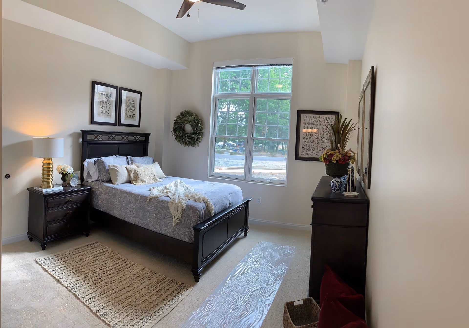 A bedroom with a large window letting in natural light. The room features a dark wooden bed with gray and white bedding, a matching nightstand with a lamp and decorative items, a dresser with plants and framed artwork on the walls. A woven rug is placed on the carpeted floor.
