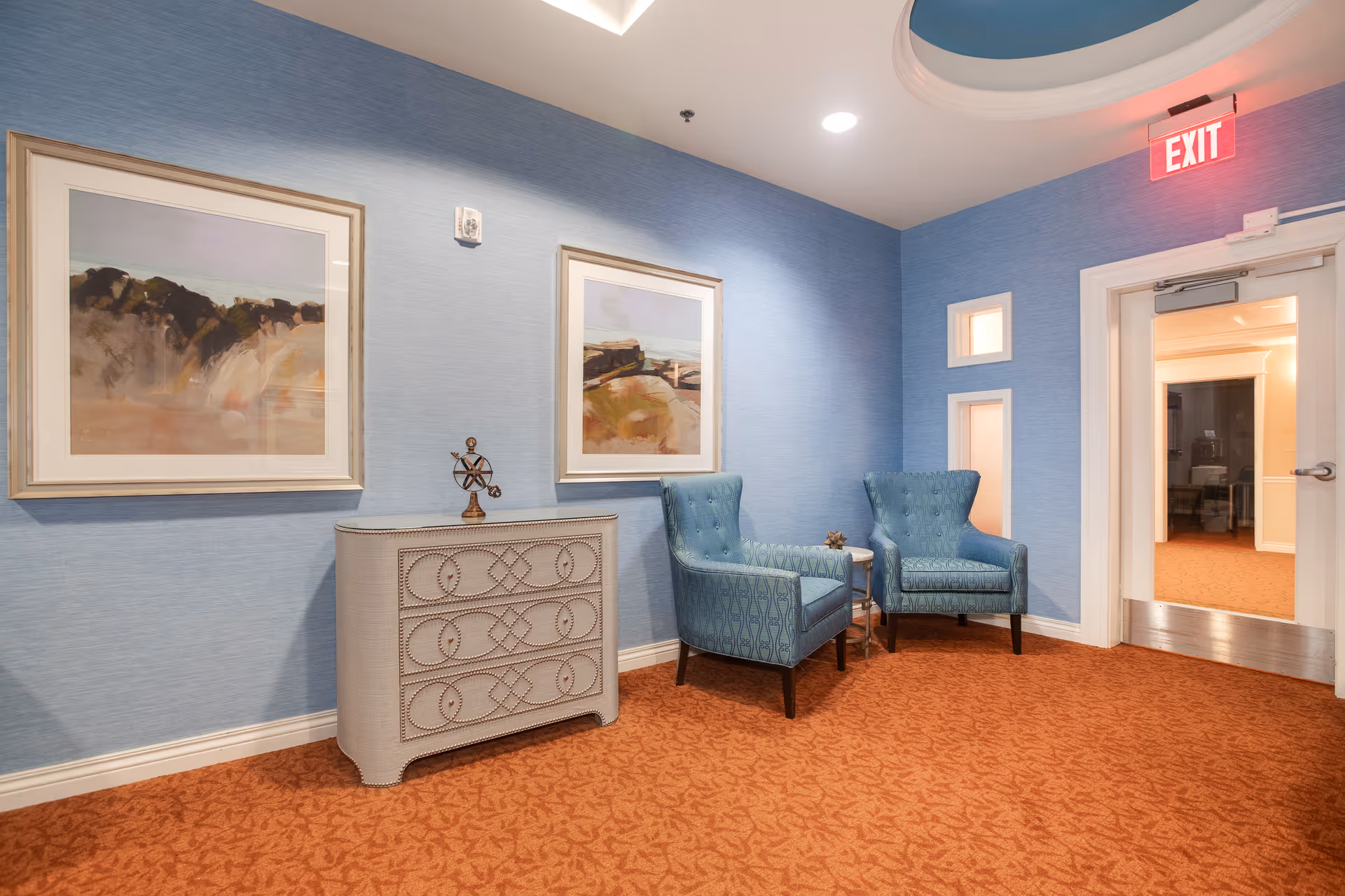 A seating area in a hallway with two blue armchairs, a decorative chest, framed artwork on blue walls, and an exit door.