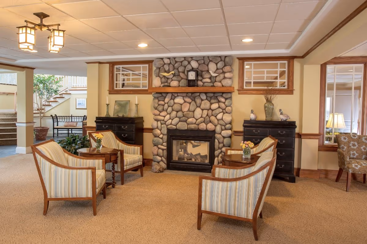A cozy seating area in a senior living facility with four striped armchairs arranged around two small wooden tables, each with a vase of flowers. The room features a stone fireplace with a clock and decorative birds on the mantel, two black dressers with decorative items, and a staircase in the background leading to another level. The walls are painted beige with wood trim, and the ceiling has recessed lighting and a hanging light fixture.