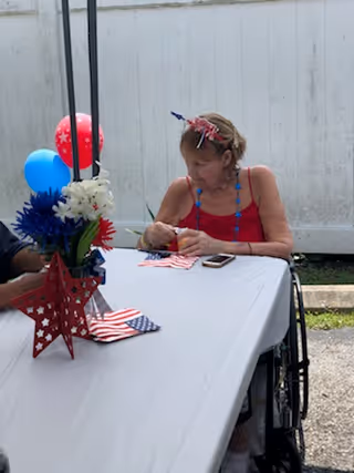 An elderly woman in a wheelchair wearing a red tank top and patriotic headband sits at a table covered with a white tablecloth. The table is decorated with red, white, and blue flowers, balloons, and star-shaped decorations. The setting appears to be outdoors with a white fence in the background.
