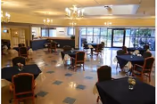 Spacious dining room with multiple tables covered in black tablecloths, each with a small floral centerpiece. Several chairs are arranged around the tables. Large windows allow natural light to fill the room, and chandeliers hang from the ceiling providing additional lighting.