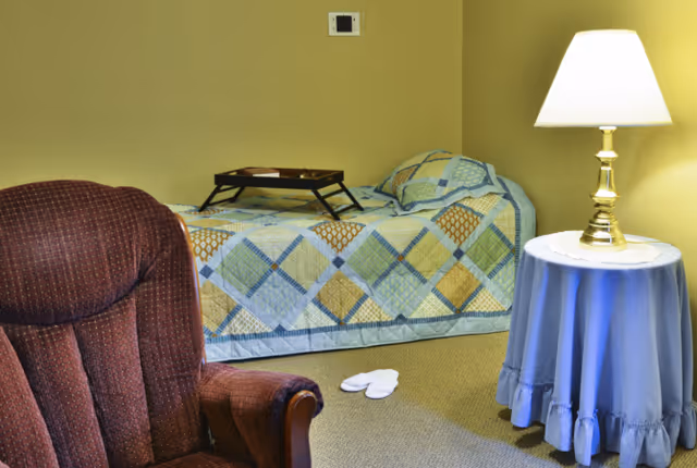 A cozy bedroom with a single bed covered in a patchwork quilt, a brown upholstered armchair, a small round table with a blue tablecloth and a lit lamp, and a pair of white slippers on the carpeted floor.