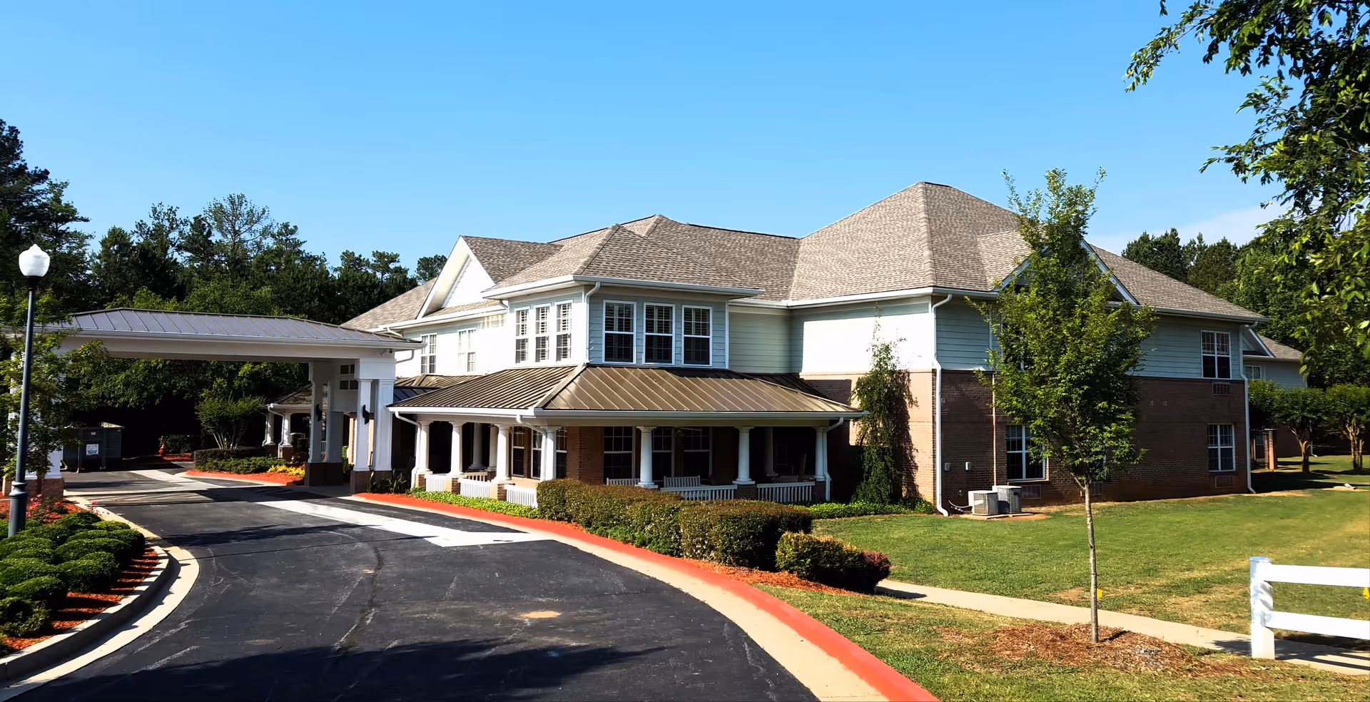 Exterior view of a two-story senior living facility building with a covered entrance driveway, surrounded by green lawns, trees, and shrubs under a clear blue sky.