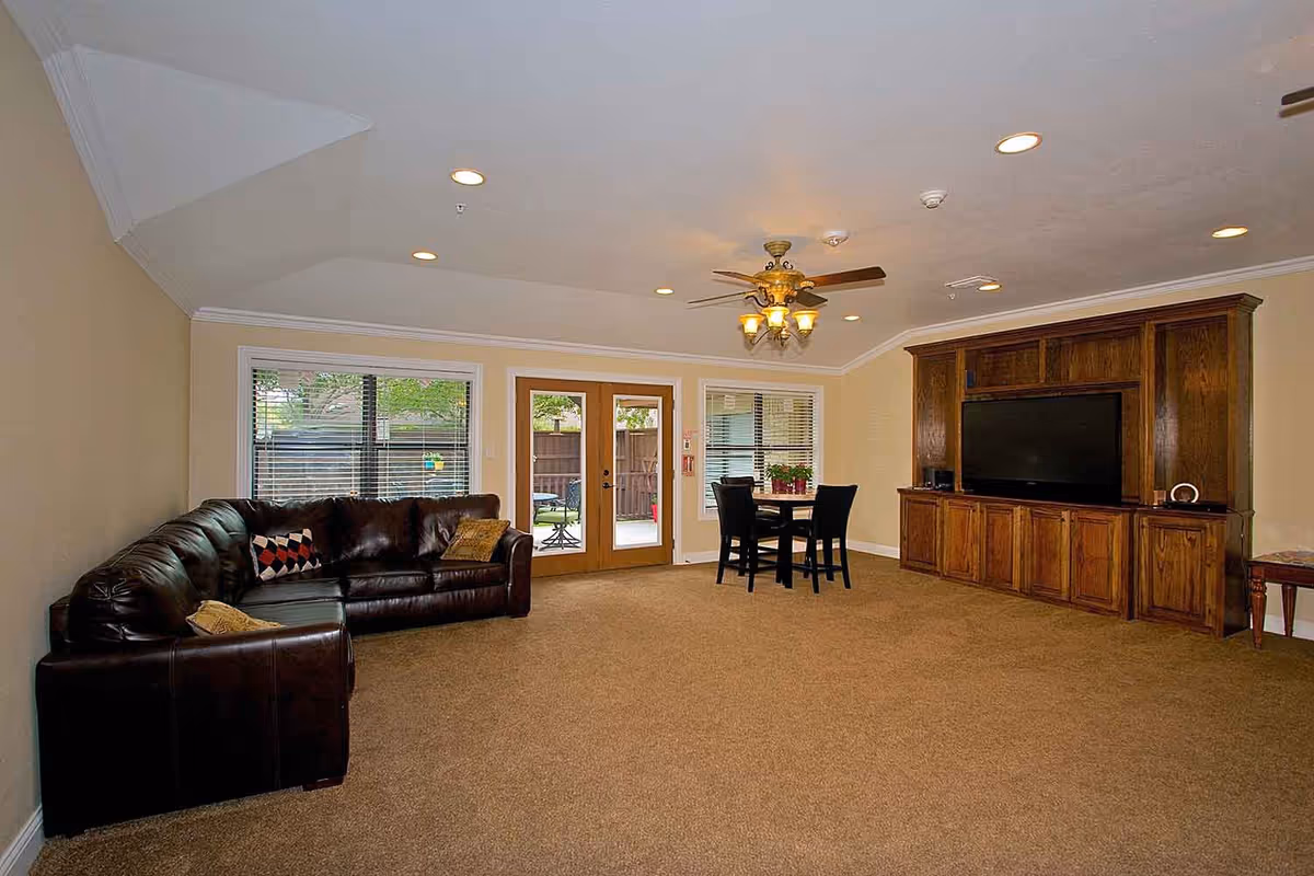 A spacious living room with beige walls and carpeted floor. There is a large dark brown leather sectional sofa with decorative pillows on the left side. In the center near the back wall, there is a small round table with four black chairs. The back wall features two large windows with blinds and a set of wooden double doors leading outside. On the right side, there is a large wooden entertainment center with a flat-screen TV. The ceiling has recessed lighting and a ceiling fan with lights.