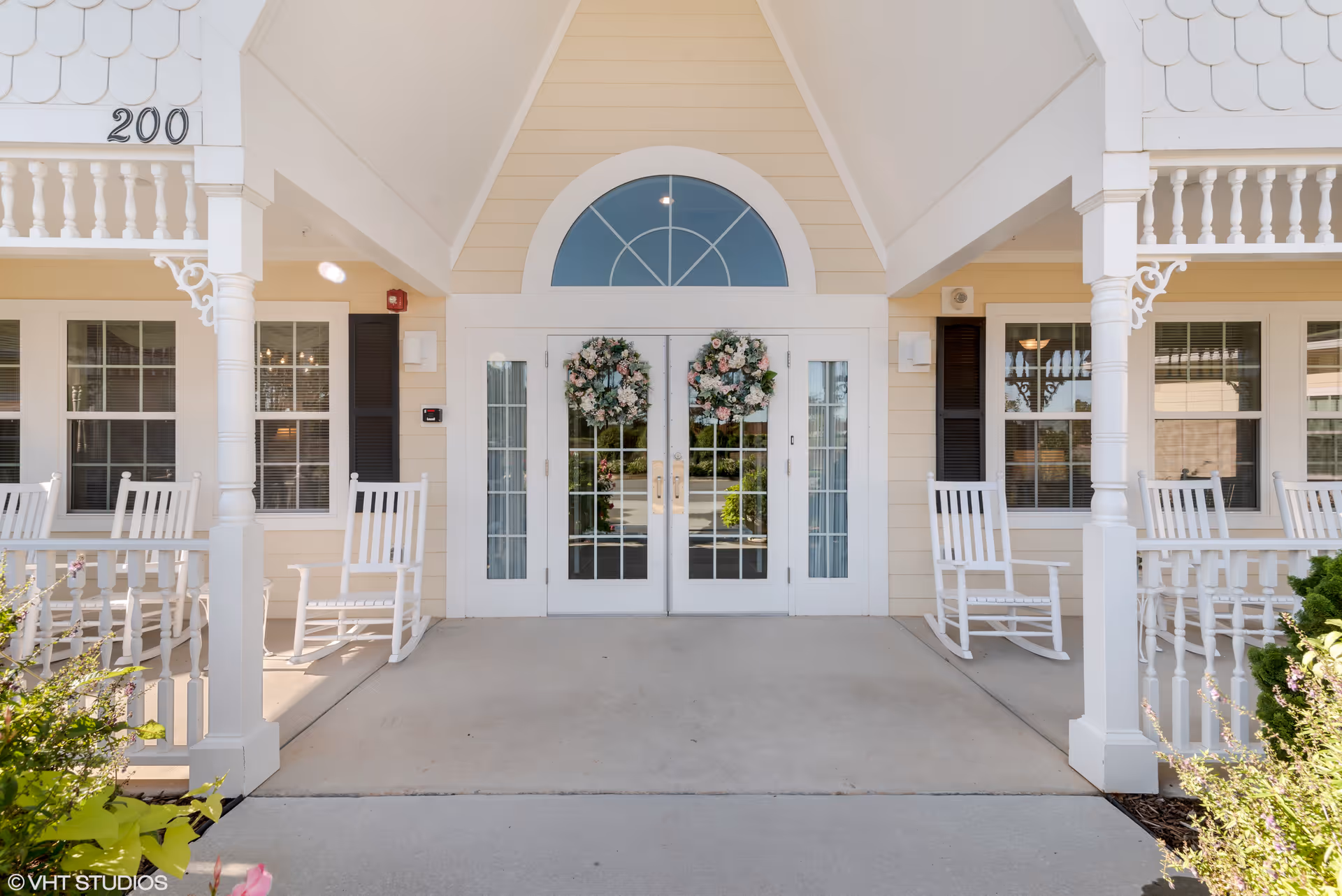 Entrance of a senior living facility with double glass doors decorated with floral wreaths. The building exterior is light yellow with white trim, featuring a covered porch with white rocking chairs on either side. The number 200 is displayed above the porch.