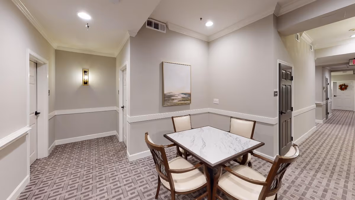 Small seating area with a marble-top table and four chairs in a carpeted hallway of a senior living facility.
