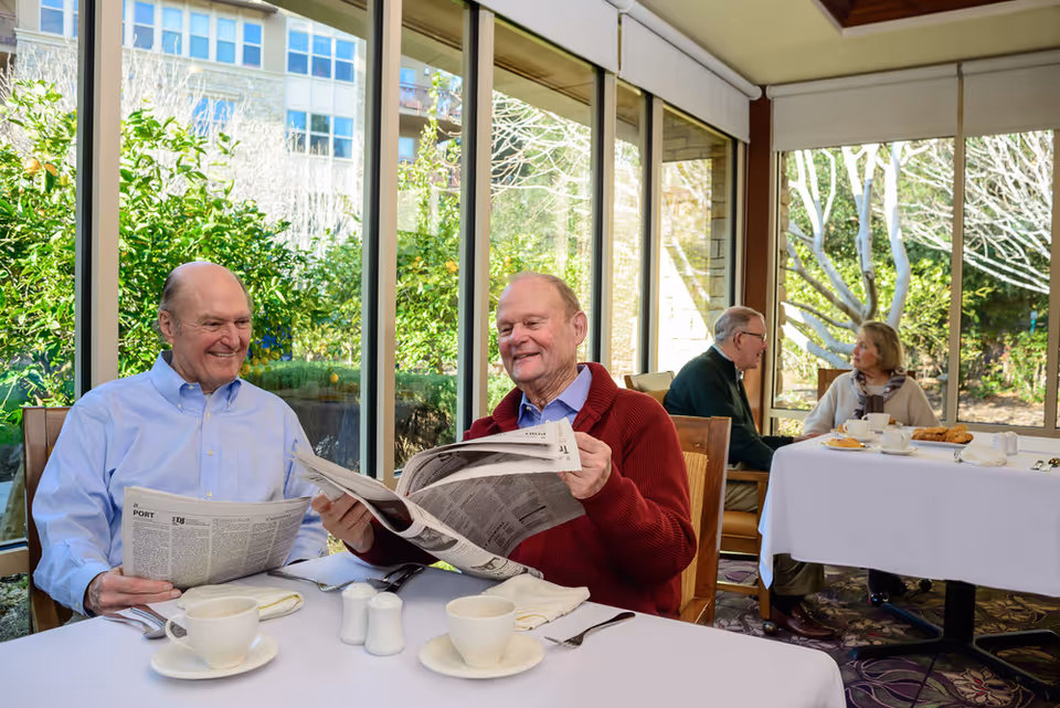 Two elderly men sitting at a dining table inside a sunlit room with large windows, reading newspapers and smiling. In the background, an elderly man and woman are seated at another table, engaged in conversation with cups and plates in front of them. Outside the windows, greenery and trees are visible.