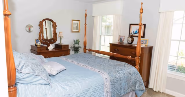 A bedroom with a wooden four-poster bed dressed in light blue bedding with a paisley pattern. There is a wooden dresser with a mirror and decorative items on top, including framed photos and a lamp. Two windows with white curtains allow natural light into the room.