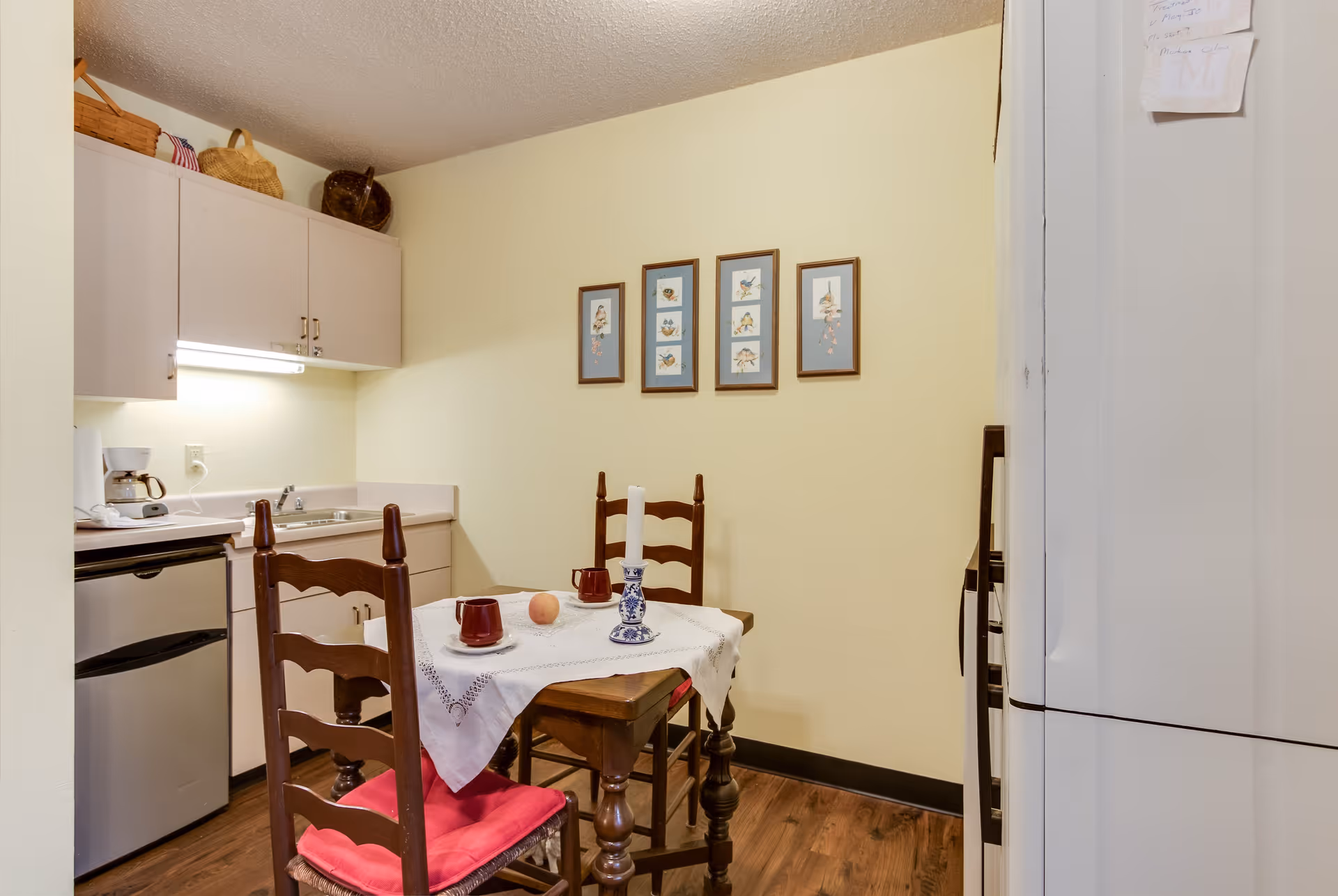 A small kitchen and dining area with a wooden table covered by a white tablecloth, two wooden chairs with red cushions, a candle holder with a white candle, two red cups, and a peach on the table. The kitchen has light-colored cabinets, a small refrigerator, a coffee maker, and a sink. Four framed pictures hang on the yellow wall above the table.