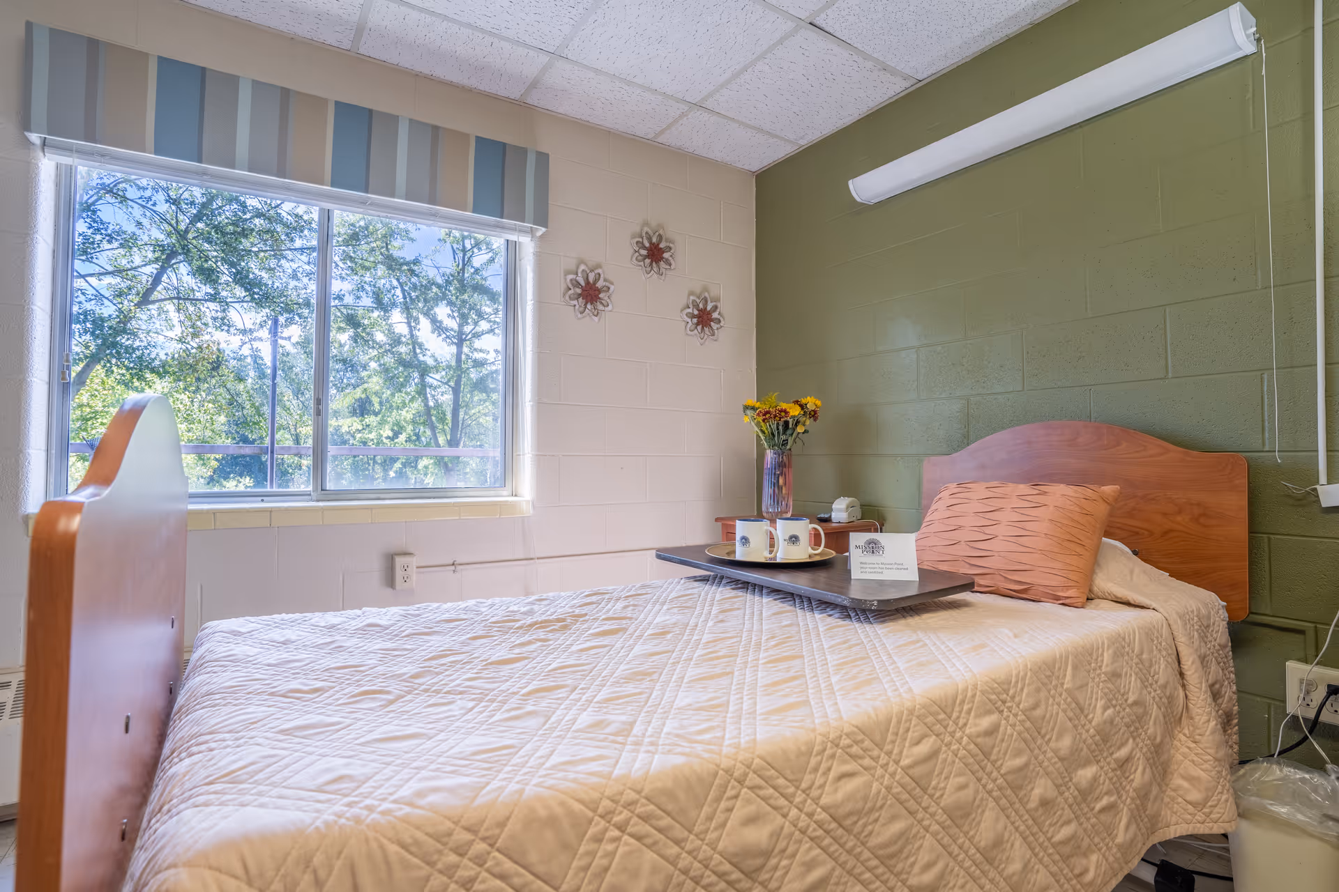 A single bed with a wooden headboard and footboard in a room with light green and beige walls. The bed is covered with a beige quilted blanket and has a peach-colored pillow. A tray on the bed holds two mugs, a small vase with flowers, and a card. There is a large window with a striped valance showing green trees outside. Three decorative flower wall hangings are on the beige wall.