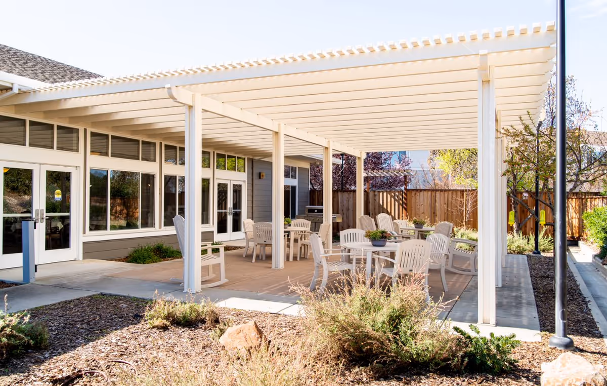 Covered outdoor patio with a white pergola and several tables and chairs outside a senior living building.