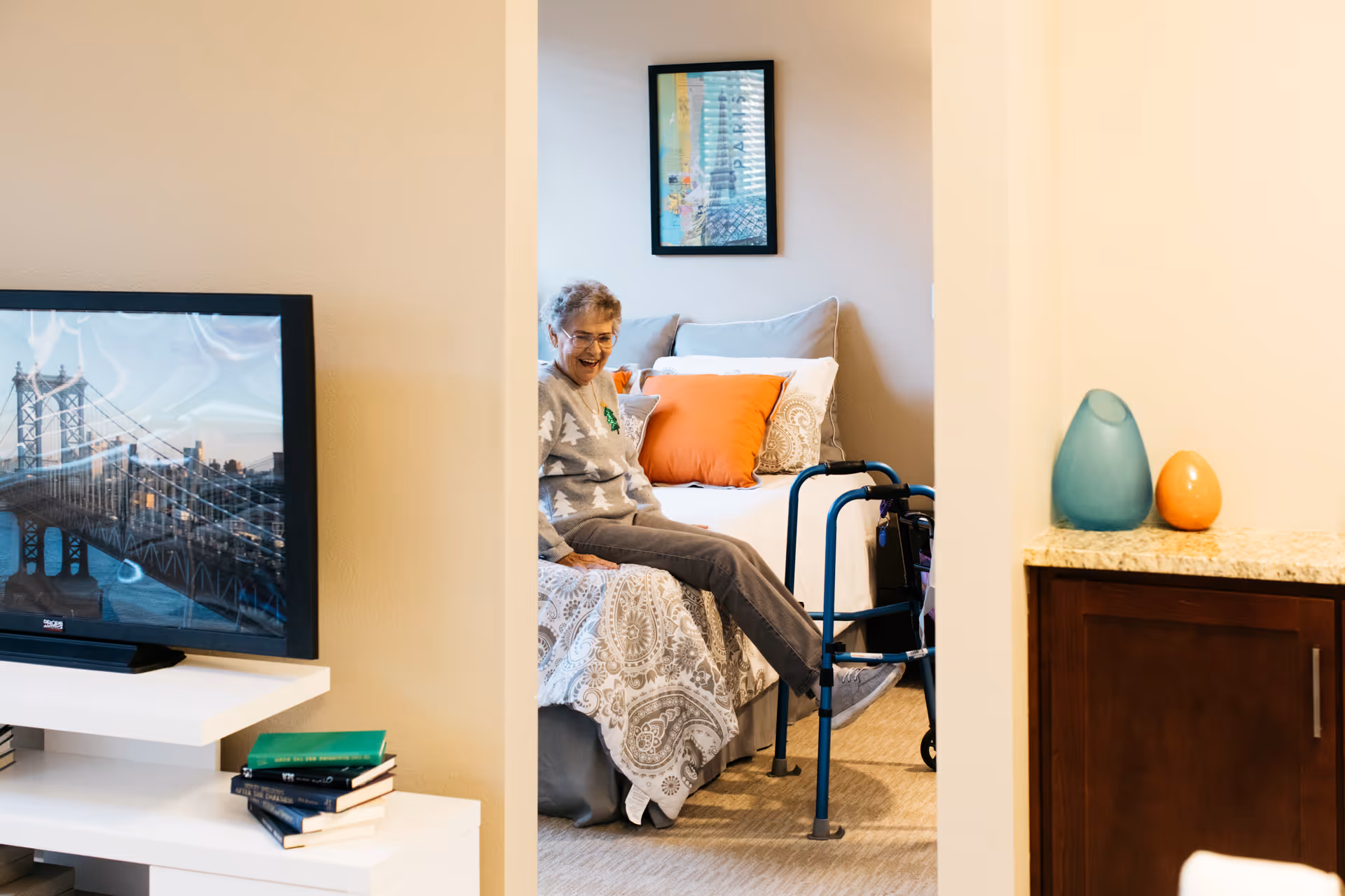 An elderly woman sits on a bed in a cheerful bedroom with a walker beside her, a TV and books on a shelf, and decorative vases on a counter.