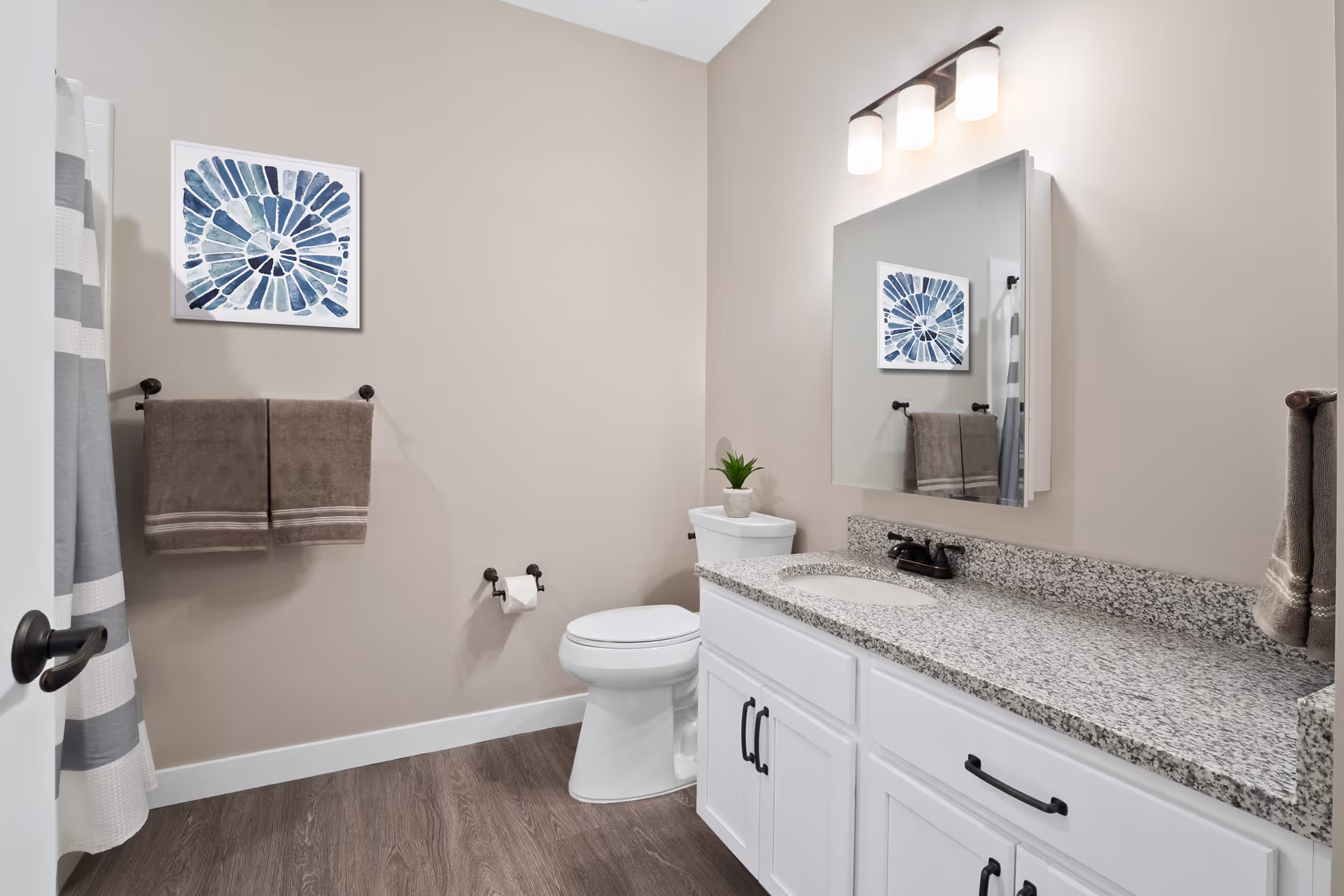 A clean and modern bathroom featuring a white toilet, a granite countertop with an under-mount sink, white cabinetry with black handles, a large mirror with three light fixtures above it, two brown towels hanging on a towel rack, a small potted plant on the toilet tank, and a framed blue and white abstract artwork on the wall. The floor has wood-like vinyl flooring and a shower curtain with gray and white stripes is partially visible.
