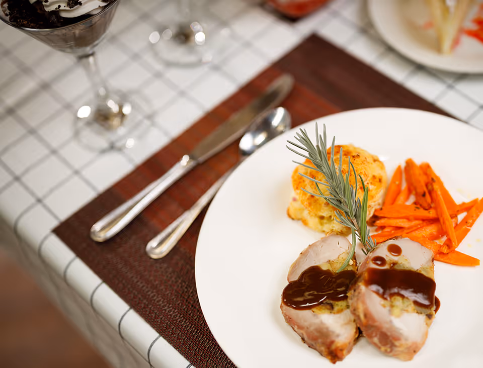 Plate with sliced meat topped with sauce, roasted carrots, a biscuit and a sprig of rosemary on a placemat with silverware.