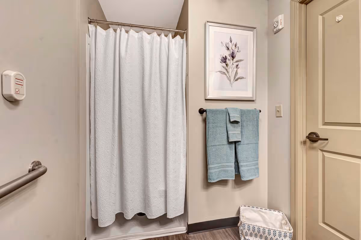 Small bathroom with a white shower curtain, teal towels on a towel rack, a grab bar, framed floral art, and a laundry basket.