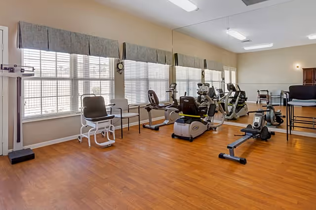 A fitness room with wooden flooring and large windows covered with blinds and valances. The room contains exercise equipment including stationary bikes, a rowing machine, and chairs. A large mirror covers one wall, reflecting the equipment and part of the room.
