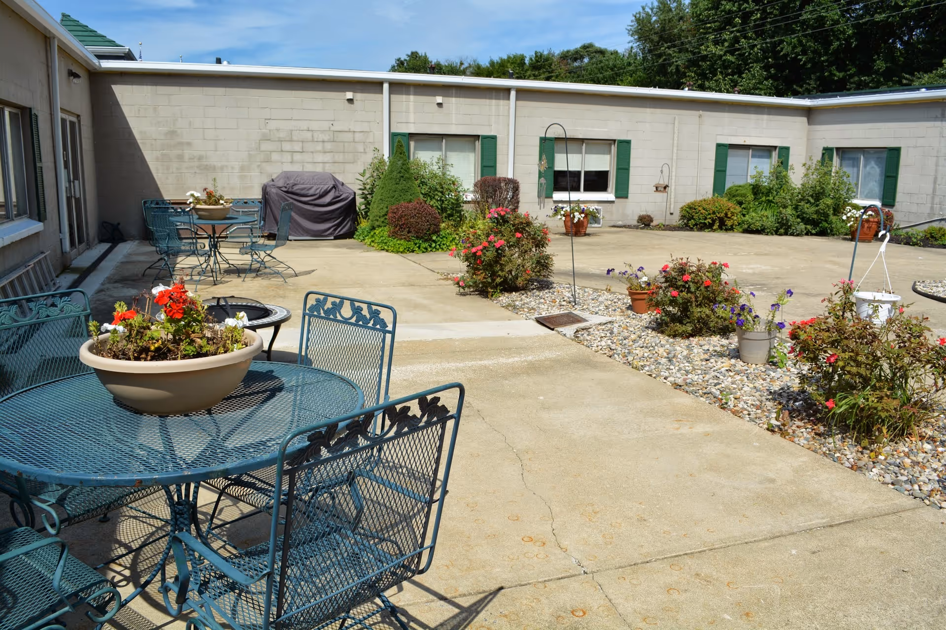 Outdoor courtyard area with metal patio tables and chairs, potted flowers on the tables, flower beds with various plants and bushes, and a building with windows and green shutters surrounding the courtyard.