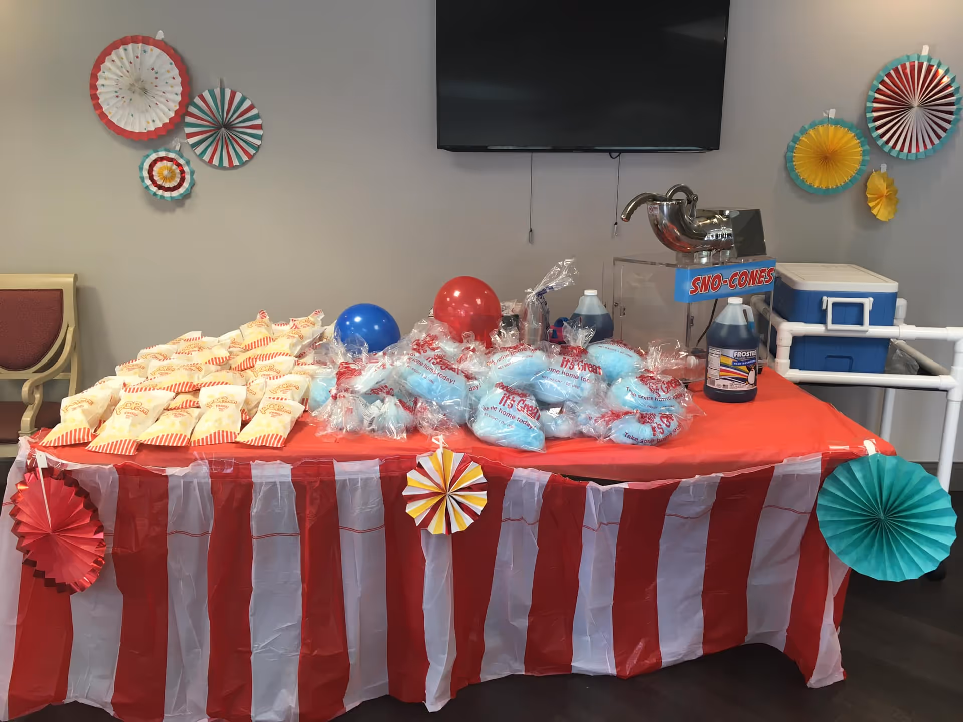 A festive table setup with a red and white striped tablecloth, decorated with colorful paper fans on the wall and table. The table holds bags of popcorn, bags of blue cotton candy, red and blue balloons, a sno-cones machine, and bottles of syrup. A TV is mounted on the wall above the table.