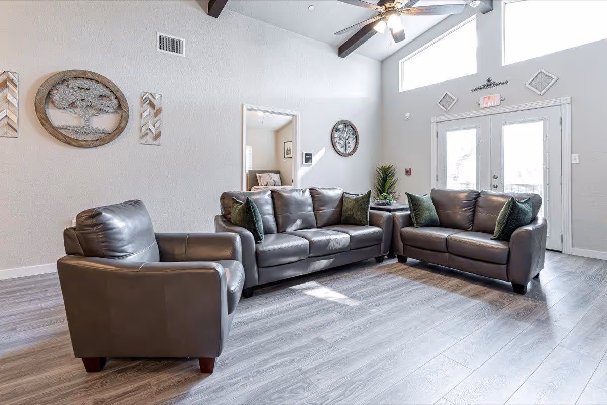 A bright living room with two gray leather sofas and one matching armchair arranged around a small side table with a plant. The room has light gray walls, wood flooring, a ceiling fan with lights, and large windows above double glass doors letting in natural light. Decorative wall art featuring tree designs is hung on the walls.
