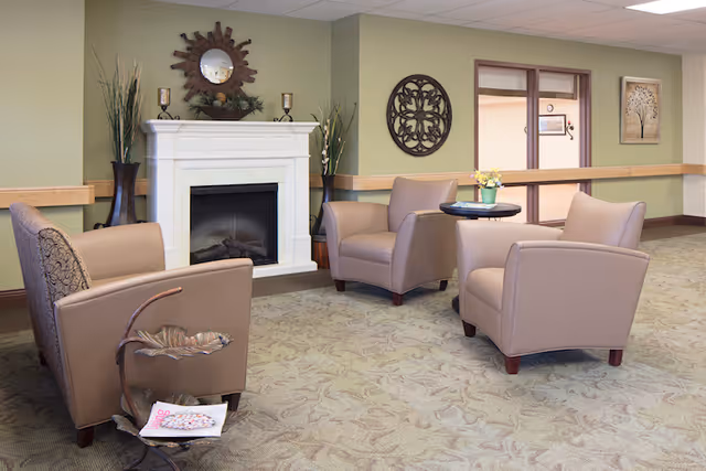 A cozy seating area with four beige armchairs arranged around a small round table with a flower pot on it. Behind the chairs is a white fireplace with decorative items on the mantel, including a round mirror and candle holders. The walls are painted light green with wooden trim, and there is a decorative wall hanging and framed artwork. The floor is carpeted with a subtle floral pattern.