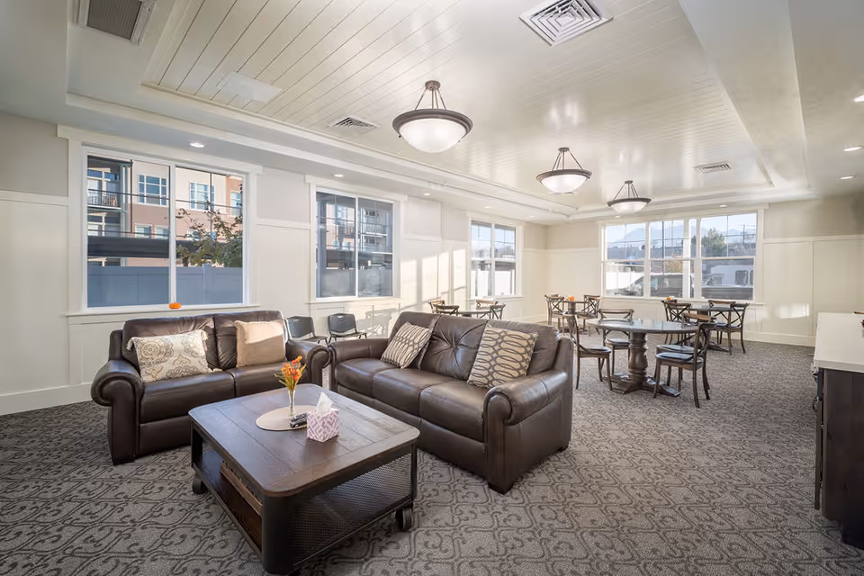 A bright and spacious senior living common area with two brown leather sofas arranged around a wooden coffee table with a small flower vase and tissue box. The room has large windows letting in natural light, patterned carpet, several round tables with chairs in the background, and ceiling light fixtures.