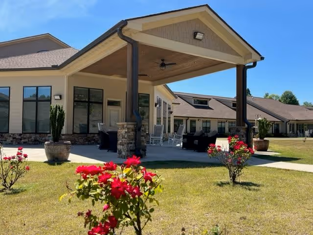 Outdoor view of a single-story assisted living facility building with a covered patio area featuring seating and rocking chairs. The foreground shows a grassy lawn with blooming red flowers and potted plants under a clear blue sky.