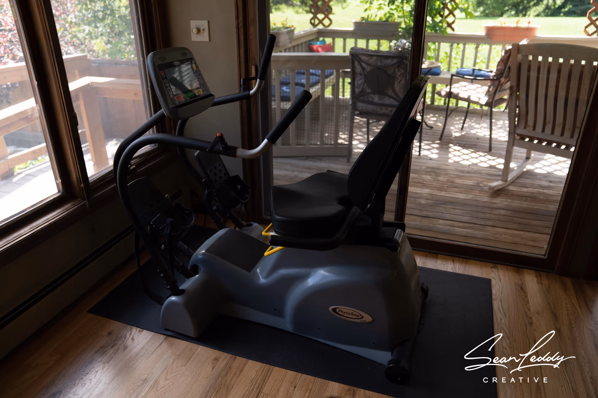A recumbent exercise bike positioned on a black mat inside a room with wooden flooring. Large windows and a glass door provide a view of an outdoor wooden deck with patio furniture and greenery beyond.