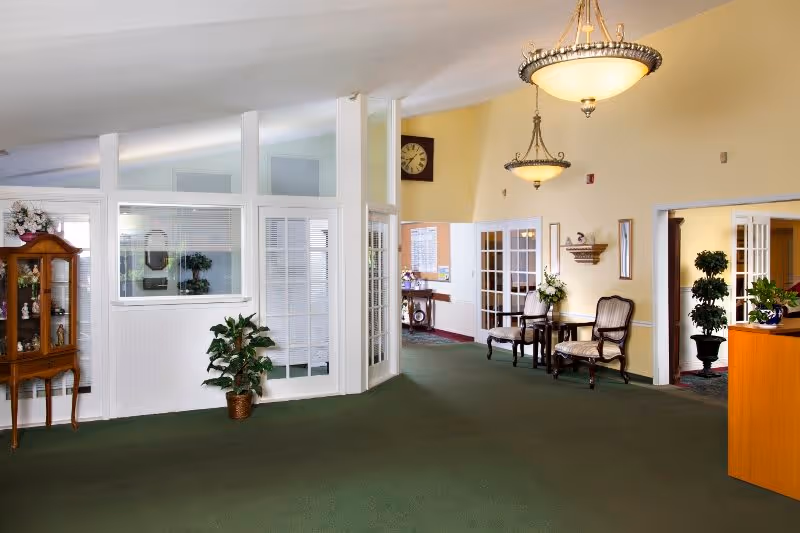 Interior view of a senior living facility lobby or common area with green carpet, cream-colored walls, two chandeliers, a wooden display cabinet with decorative items, potted plants, and seating area with two chairs and a small table. There are glass-paneled doors and windows, and a large clock on the wall.