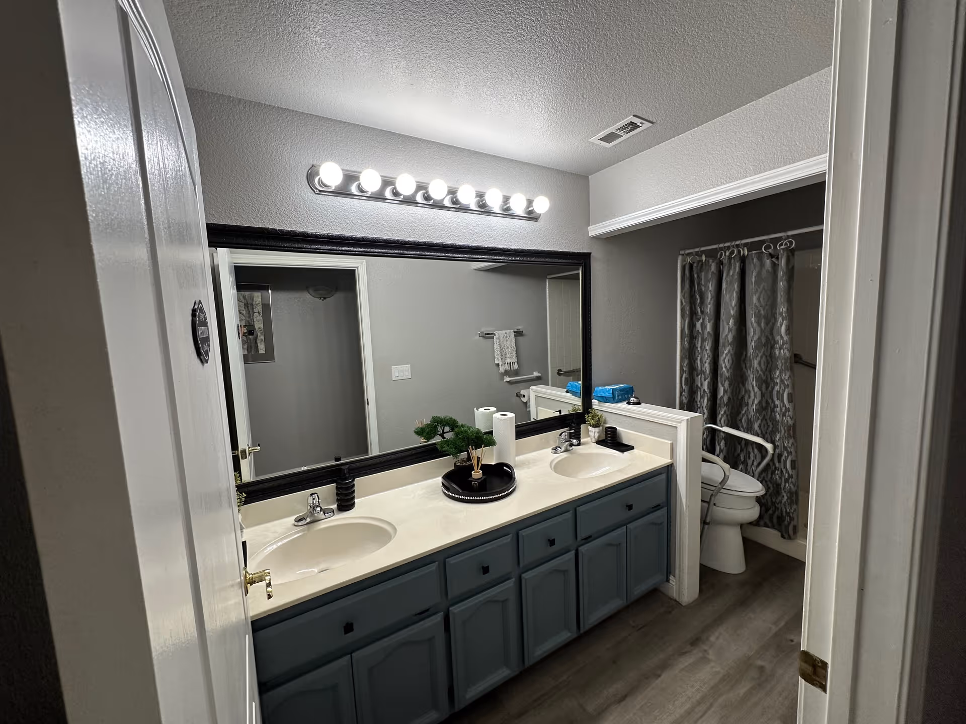 Interior view of a bathroom with a double sink vanity featuring blue cabinets and a large mirror above. There is a row of bright vanity lights above the mirror. To the right, there is a toilet with a grab bar and a shower with a patterned curtain. The floor is wood-style laminate.
