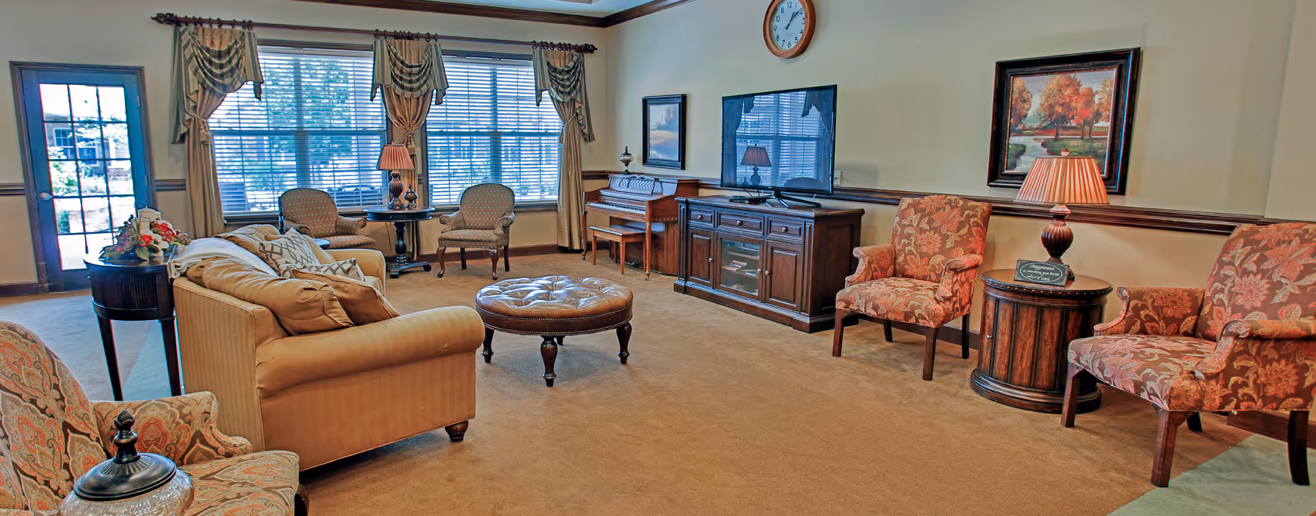 A cozy living room with beige carpet and walls, featuring a beige sofa, patterned armchairs, a round tufted ottoman, a wooden TV stand with a flat-screen TV, a piano, and large windows with curtains letting in natural light. There are also table lamps and framed artwork on the walls.