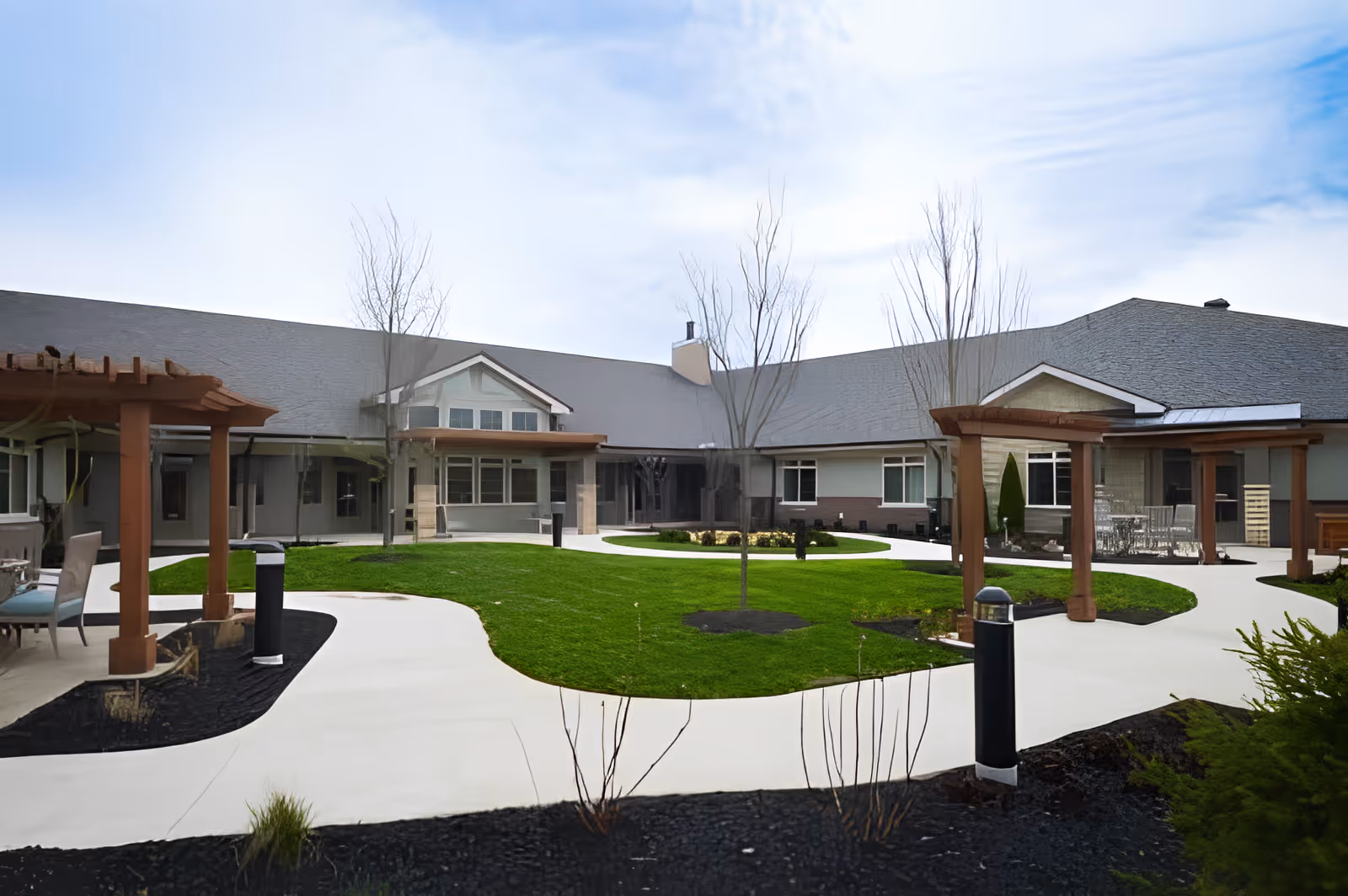 Outdoor courtyard area of The Ganzhorn Suites featuring a well-maintained green lawn, paved walkways, leafless trees, and wooden pergolas with seating areas. The building surrounds the courtyard with multiple windows and a gray roof under a partly cloudy sky.
