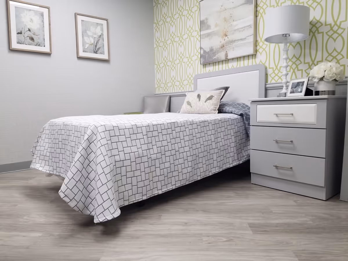 A neatly made single bed with a white and gray patterned bedspread in a room with light wood flooring. The bed has a gray and white headboard and two pillows, one with a leaf design. Next to the bed is a gray nightstand with three drawers, a white table lamp, a framed photo, and a vase of white flowers. The walls are decorated with two framed floral prints and a larger abstract painting on a green patterned accent wall.