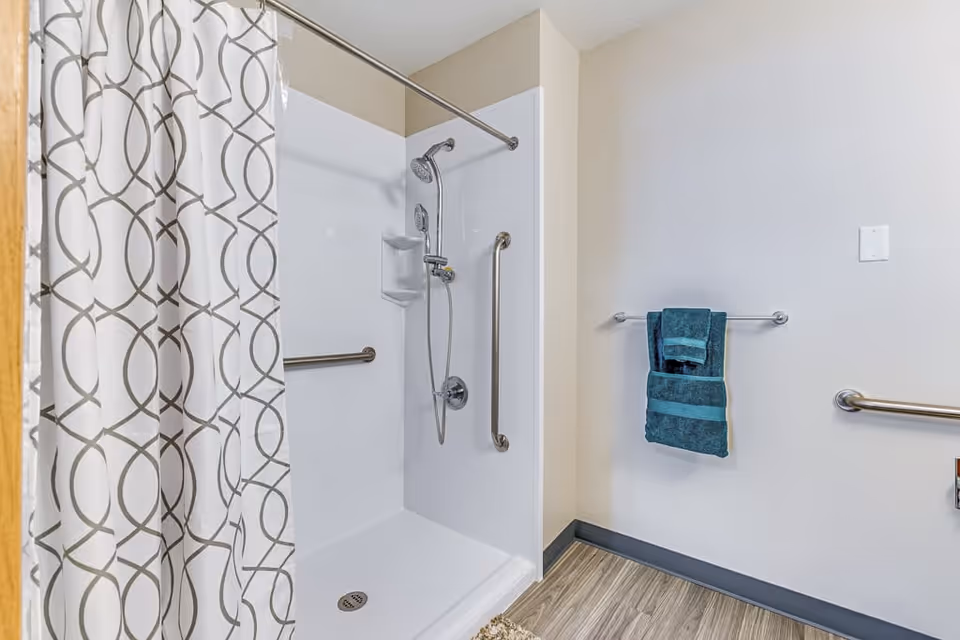 A bathroom shower area with a white shower stall, a silver showerhead, and two grab bars for support. There is a white shower curtain with a gray circular pattern partially drawn to the left. On the right wall, there is a towel rack holding two teal towels. The floor has wood-like vinyl flooring.