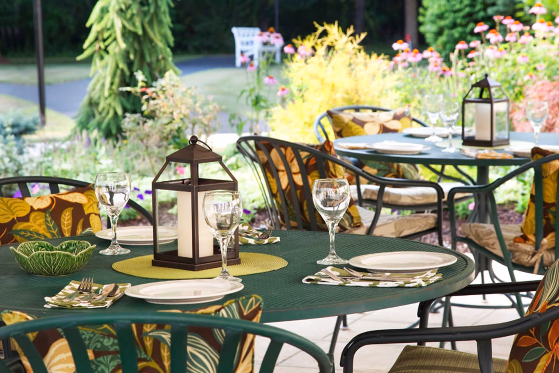 Outdoor patio dining area with green metal tables and chairs adorned with colorful leaf-patterned cushions. Tables are set with white plates, wine glasses, napkins, and decorative lanterns. Surrounding the patio are vibrant flowers and greenery.