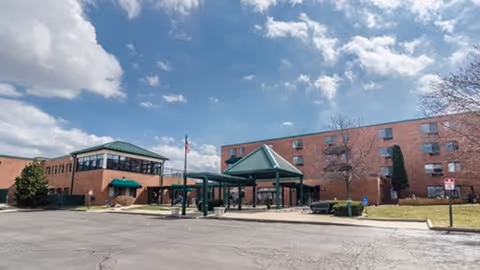 Exterior view of a senior living facility with a large brick building, green roofs, and a covered entrance area under a partly cloudy sky.