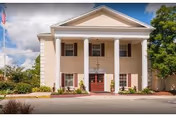 Front exterior view of a two-story building with four white columns supporting a triangular pediment above the entrance. The building is beige with red doors and windows, surrounded by greenery and a clear sky with some clouds.