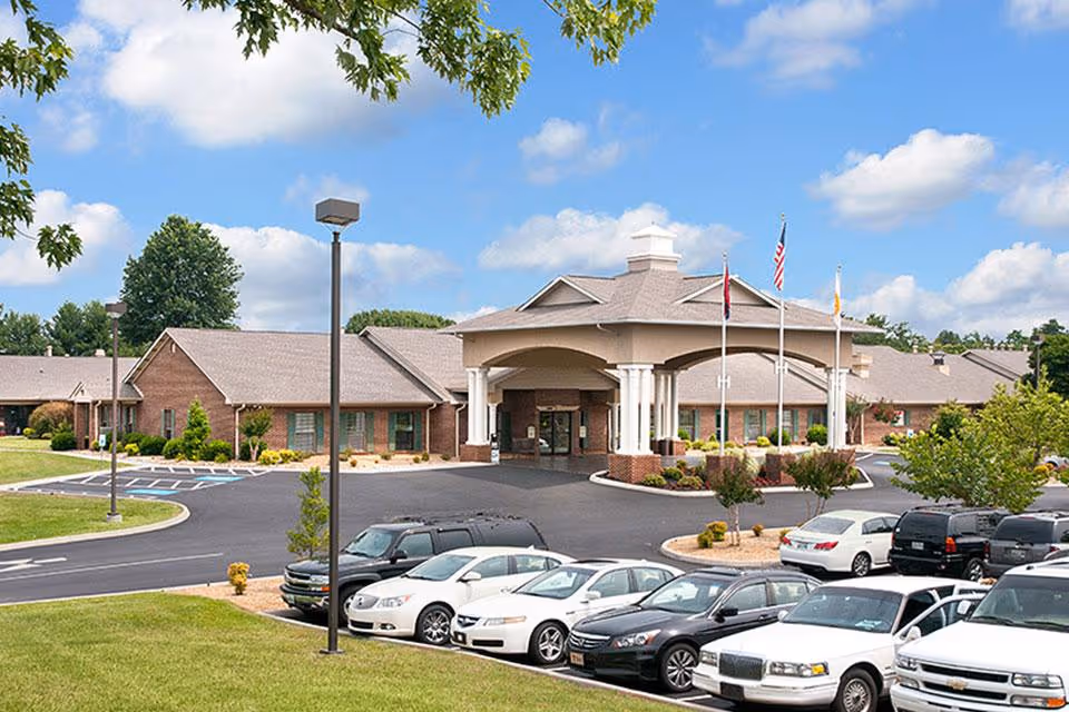 Front exterior of The Heritage Center, a single-story brick senior living facility with a covered main entrance, flagpoles, parked cars and landscaped grounds under a blue sky.