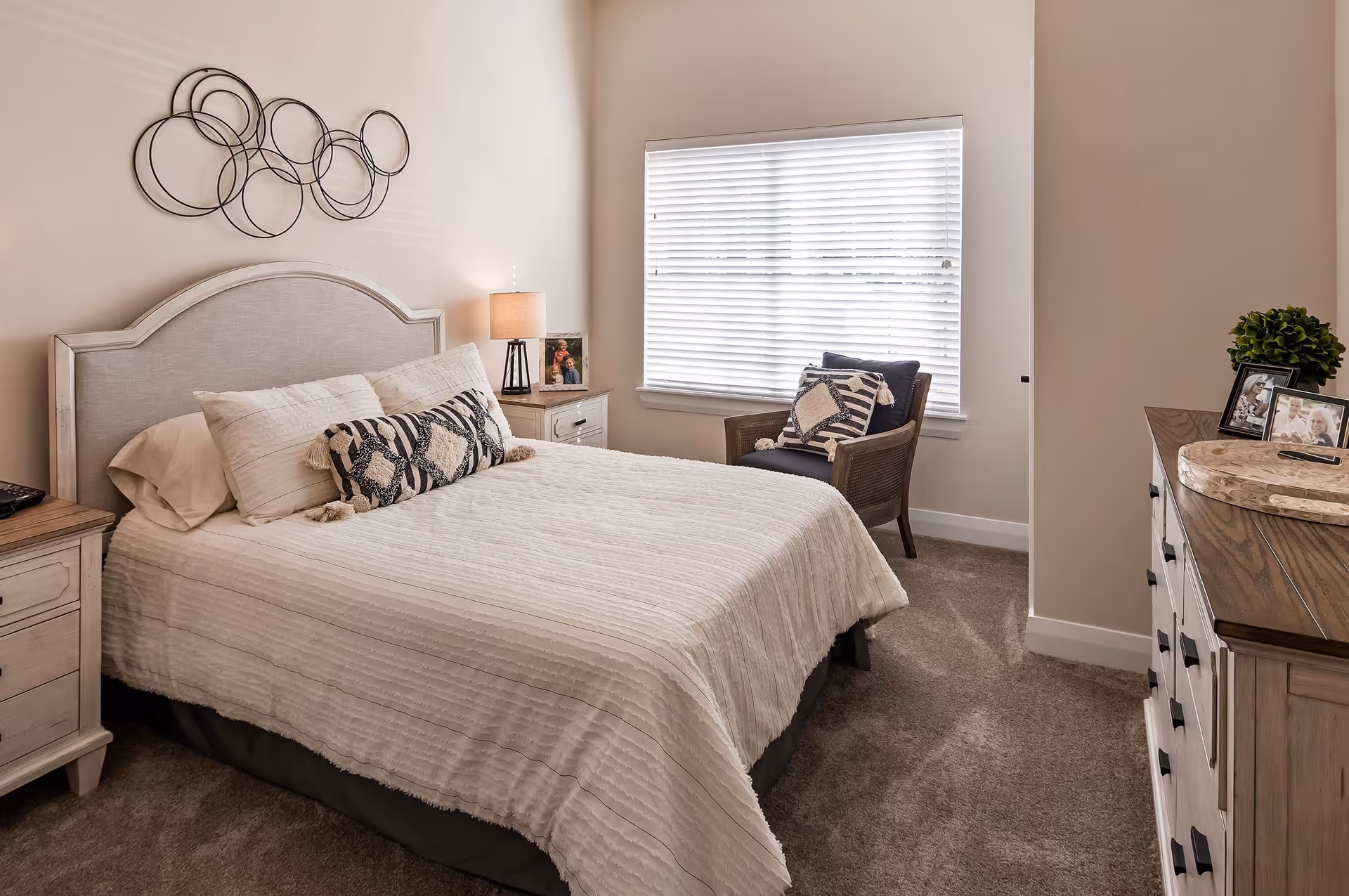 A cozy bedroom with a neatly made bed featuring white bedding and decorative pillows. There is a nightstand with a lamp and a framed photo on one side of the bed. A window with white blinds lets in natural light. A cushioned chair with patterned pillows is placed near the window. Across from the bed is a wooden dresser with framed photos and a decorative tray on top. The walls are painted a soft beige color, and a modern circular metal wall art hangs above the bed.