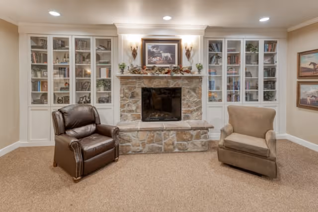 A cozy living room area with a stone fireplace in the center, flanked by built-in white bookshelves filled with books and decorative items. Two armchairs, one brown leather and one beige fabric, are positioned facing the fireplace. Above the fireplace is a framed painting of dogs, and two wall sconces provide warm lighting. The room has beige walls and carpeted flooring.