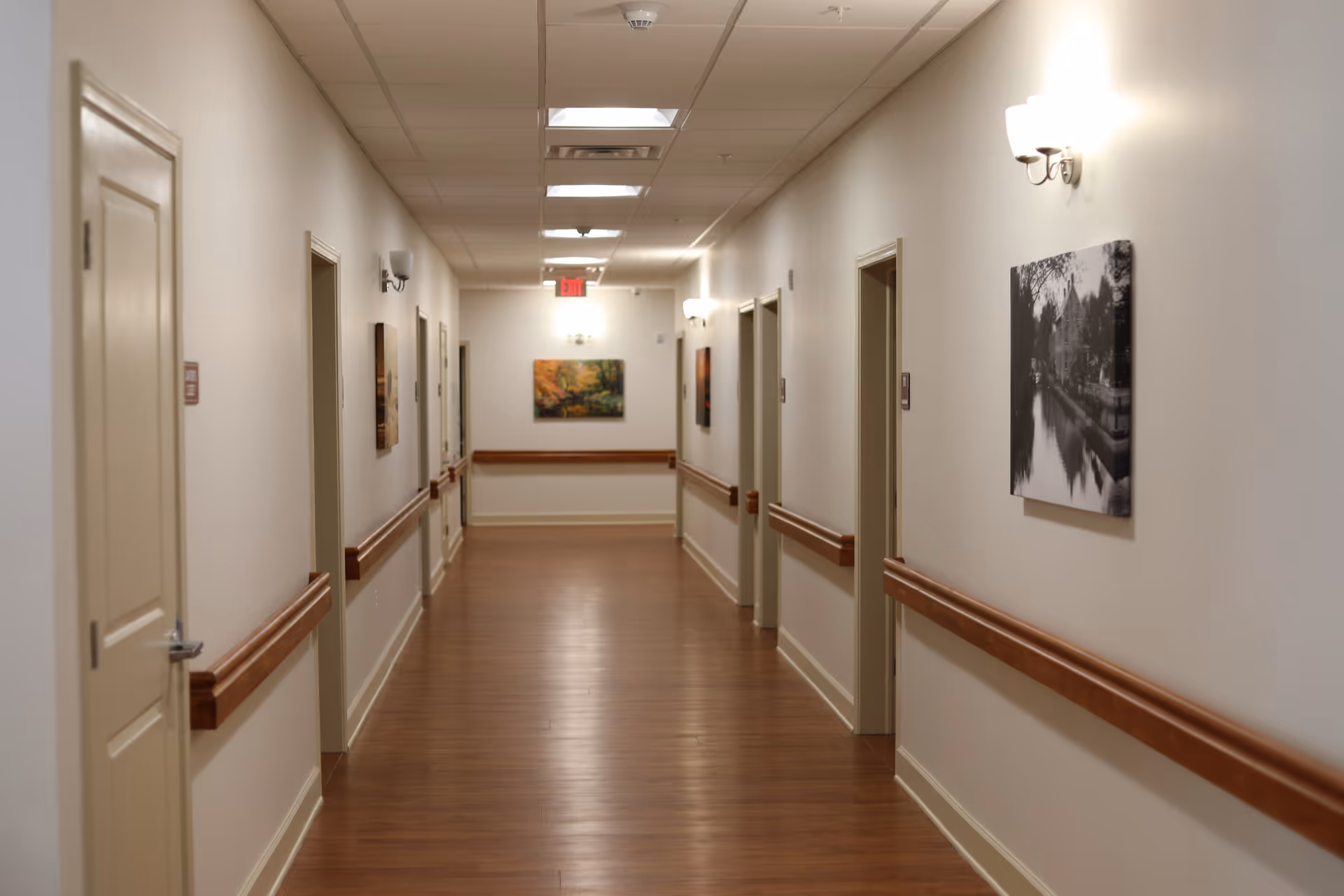 A clean, well-lit hallway in a senior living facility with wooden floors, beige walls, handrails on both sides, several closed doors, and framed artwork hanging on the walls.