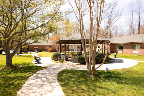 Outdoor courtyard area at Autumn Care of Saluda Center featuring a paved walking path, green grass, trees, benches, and a covered gazebo structure surrounded by a single-story brick building.