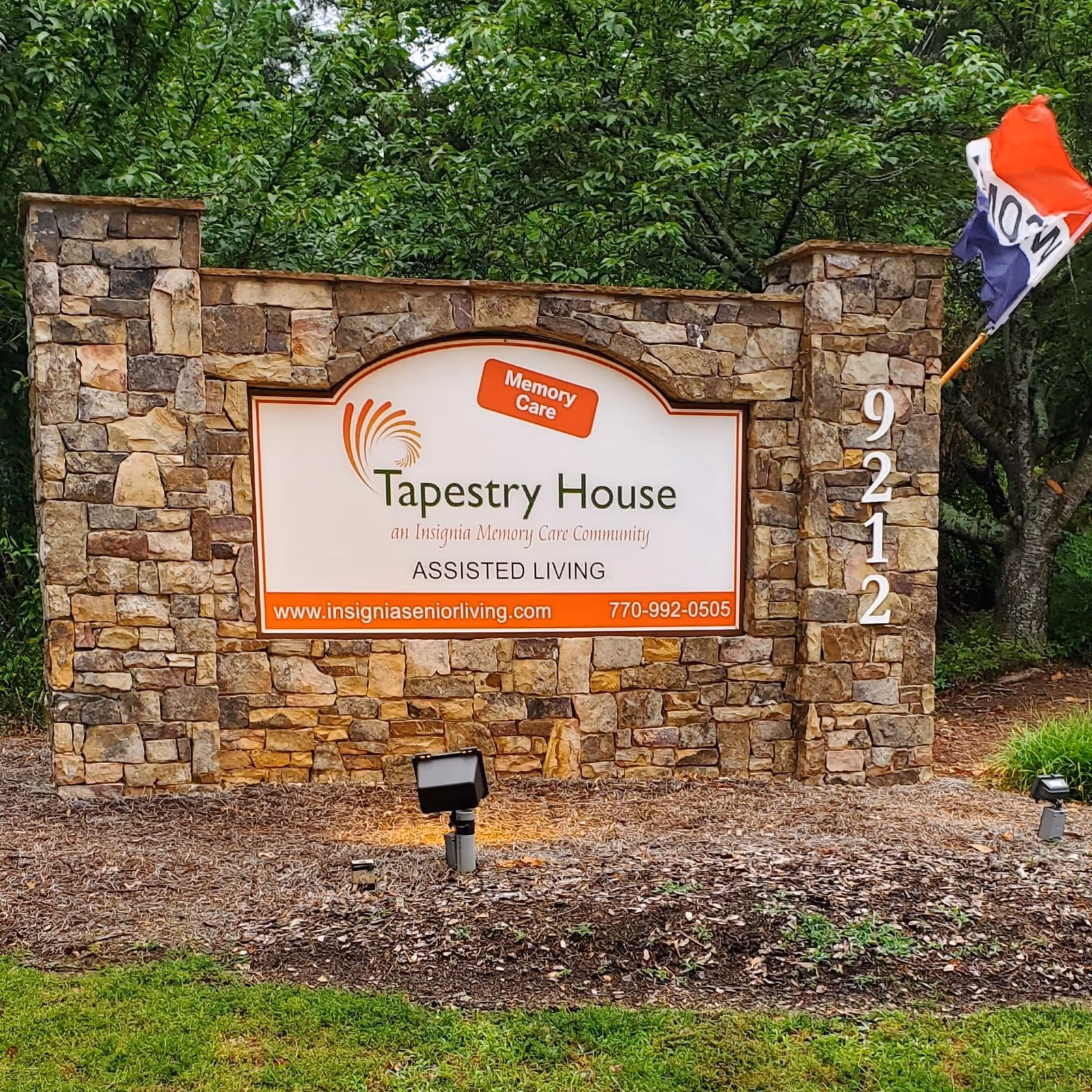 Stone sign for Tapestry House Memory Care at Alpharetta with the address number 9212 on the right side and a flag on a pole behind the right pillar. The sign includes the text 'Memory Care', 'Tapestry House an Insignia Memory Care Community', 'ASSISTED LIVING', and the website www.insigniaseniorliving.com with phone number 770-992-0505.