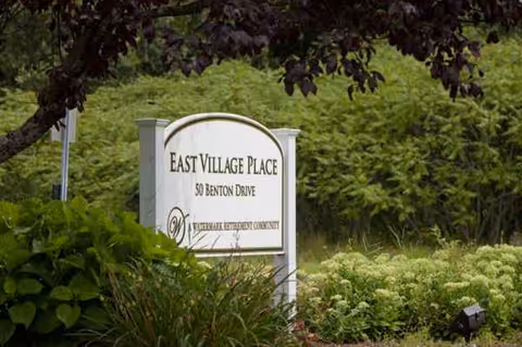 A sign reading East Village Place sits among lush landscaping at the facility entrance.
